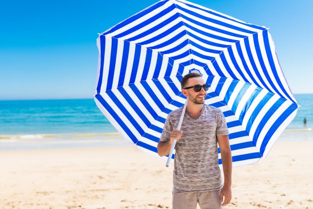Young man in sunglasses walking on the beach with sun solar umbrella on sea resort. Summer vocation.